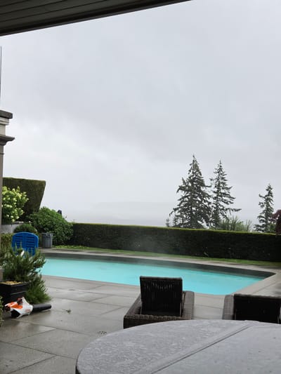 An overcast day in West Vancouver, Canada, shows a backyard scene with a swimming pool. The pool is a bright turquoise blue, surrounded by a grey concrete patio. On the patio, a blue plastic chair and a grey trash can are visible to the left of the pool. To the left of the pool, a small evergreen plant in a black pot sits next to a white and orange leaf blower, with the brand "STIHL" visible on it. Two dark brown wicker lounge chairs face the pool, and a portion of a large grey outdoor table is in the foreground, with water droplets on its surface. A neatly trimmed green hedge runs along the edge of the property, with several tall evergreen trees visible beyond it. The sky is a uniform grey, indicating a cloudy or rainy day.