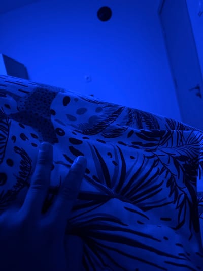 The scene depicts an indoor environment, likely a bedroom, in Nadarevo, Bulgaria, characterized by a pervasive blue ambient light. A human left hand, with fingers slightly curled, is prominently visible in the lower-left to mid-foreground, resting upon a patterned fabric. This fabric, likely a duvet cover or blanket, displays a motif of large tropical leaves and irregular spots in varying shades of blue and darker blue/black under the present lighting conditions. The fabric appears loosely draped. The upper portion of the image reveals a flat, light-colored ceiling with a dark, circular object mounted centrally, possibly a recessed light fixture or sensor. A section of a wall with a discernible wall fixture, potentially an electrical outlet, is visible towards the upper-right. No other individuals or specific actions are evident.