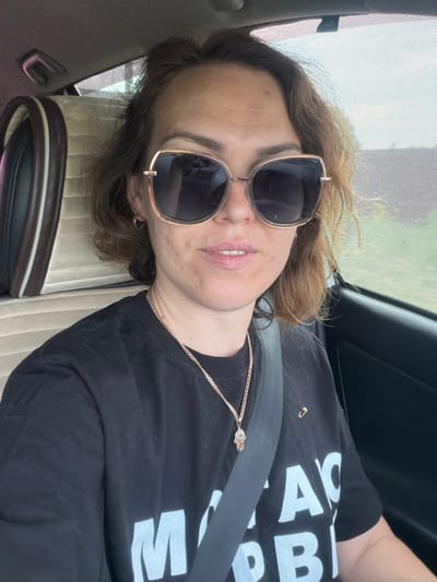 A woman is sitting in the passenger seat of a car. She is wearing sunglasses, a black t-shirt with white lettering, and a gold necklace. The car's interior is visible, including a headrest and a seatbelt. The window is open, and a blurry green landscape is visible outside. The image is a selfie, taken by the woman. The location is the city of Subkhankulovo, Russia.