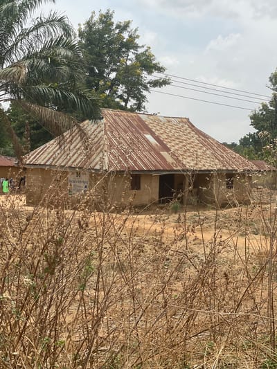 This full-shot image, taken in Unguwar Gaya, Nigeria, depicts a rustic outdoor scene centered around a modest building. The structure features a corrugated metal roof, heavily rusted with a distinctive brown and orange pattern, suggesting age and exposure to the elements. The walls appear to be constructed from earth or concrete, displaying a weathered texture, and include small, dark window openings and a recessed entrance.
The foreground and surrounding areas are characterized by dense, dry, brown vegetation, including withered bushes, weeds, and grasses, indicative of a dry climate or unkempt land. A dirt path or road runs alongside the building. In the background and to the left, several trees are visible, including a large tree with green foliage and yellow blossoms, and a palm tree with long fronds. Overhead, the sky is overcast with light grey clouds, and multiple power lines stretch across the upper part of the frame.
A white and blue sign or poster is affixed to the wall of the building. Although partly obscured and blurry, the text "MARCHING FOR BETTER Nigeria" and a number "207" are discernible. No people are visible in the image, and no specific activity or event is occurring. The scene appears to be set during daylight hours under a cloudy sky, conveying a sense of quietude in a rural or semi-rural setting.