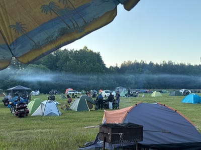This image depicts an outdoor camping scene, likely a motorcycle rally, set in a grassy field within the Валожынскі сельскі Савет region of Belarus. In the foreground, a large patterned umbrella with a palm tree motif partially obscures the view. The middle ground is populated with numerous tents of various colors and sizes, suggesting an organized event. Several motorcycles are parked amongst the tents, some appearing to be custom or touring models. People are visible interacting around the tents and vehicles, with some standing and others seated. In the background, a line of trees forms the horizon, with a light haze or mist rising in the middle distance. Several cars are also present, interspersed with the camping setup, indicating attendees arrived by various means. The overall atmosphere appears to be relaxed and communal, typical of an outdoor gathering or festival. The sky is clear and blue, suggesting daytime conditions, possibly late afternoon or early evening due to the soft light.
