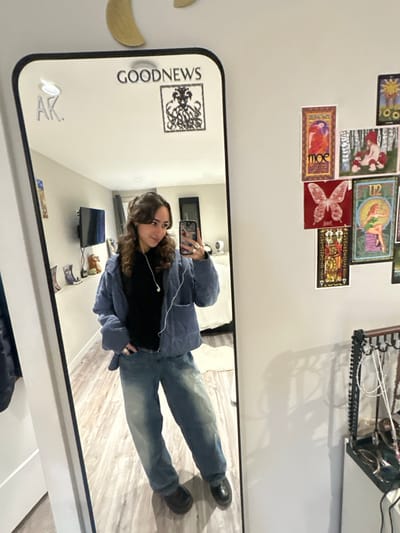 A mirror selfie shows a woman wearing blue jeans, a black top, and a light blue quilted jacket. She holds her phone up to the mirror, capturing her reflection. The room in the mirror features a TV, while the mirror itself has "GOODNEWS" printed on it and is decorated with a sun, flowers, and other assorted cards.