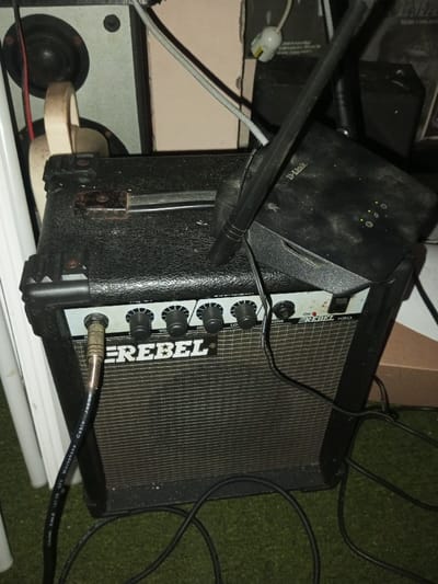 A compact "REBEL" brand guitar amplifier sits prominently in the frame. The amplifier features several knobs for volume and tone adjustment, a microphone input, and a power switch. A D-Link wireless router sits on top of the amplifier. Cords from the guitar and the router create a tangled mess in the foreground. The amplifier and router are placed in front of a larger speaker and are positioned on a surface covered in green, artificial turf.