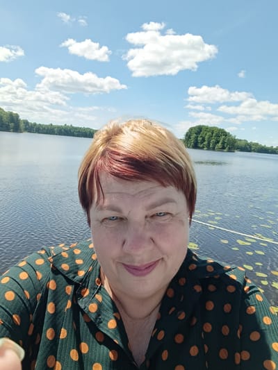 A close-up selfie shows a woman with blond and auburn hair, blue eyes, and a smile. She is wearing a dark green shirt with orange polka dots. The background features a calm lake under a bright blue sky with fluffy white clouds. The shoreline in the distance is lined with trees. Lily pads are visible on the surface of the water in the foreground, with a white rope also visible. The location is specified as Мяжанскі сельскі Савет, Belarus.