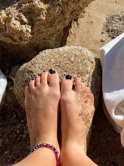 The image presents a close-up, downward-facing view of a person's bare feet in a sandy, rocky coastal environment within Megalochori, Greece. Both feet feature dark, likely black, nail polish on the toes. The right foot is partially covered with fine sand, particularly on the upper surface and between the toes. The left foot is cleaner, with a red string bracelet containing blue and white 'evil eye' beads worn around the ankle. The feet are positioned on a sand-covered, light-colored rock in the foreground. Surrounding elements include fine-grained sand, small pebbles, and larger, irregular tan-colored rock formations, suggesting a beach or rocky shoreline. A segment of a white rolled towel or fabric is visible on the sand to the right, and a white plastic bag or object is partially discernible on the far left. The scene is illuminated by bright sunlight, casting distinct shadows.