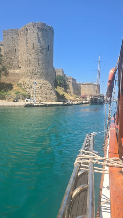 The image captures a view from a boat sailing in the turquoise waters of Kyrenia Harbor, Northern Cyprus. On the left, a section of the medieval Kyrenia Castle walls, constructed from large, rough-hewn stones, dominates the scene. The castle's massive cylindrical tower and adjoining walls rise above a grassy embankment. A light-colored car is parked on a paved area along the harbor's edge. In the middle ground, a larger wooden sailing ship with masts is moored in the harbor. To the right, the foreground shows the railing and wooden deck of the boat from which the photo is taken, with ropes secured to the metal railing. The sky is a clear, vibrant blue.
