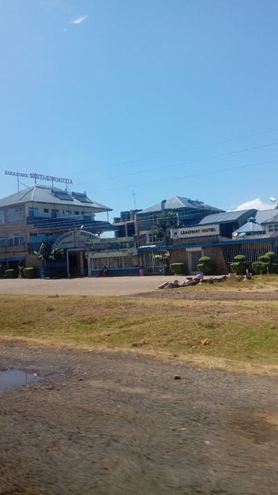 The image shows Leadway Hotel, a multi-story building under a clear blue sky. The hotel's signage is prominently displayed. A grassy lawn and a paved area border the hotel's entrance. The foreground reveals a rough, dirt road with a small puddle. Power lines stretch across the sky above the building.