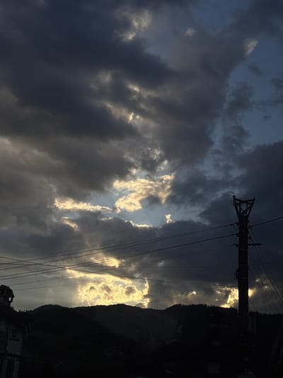 The image shows a view of the sky over Borino, Bulgaria. The sky is filled with dark, ominous clouds, but there are a few breaks in the clouds that allow sunlight to shine through. In the foreground, there is a telephone pole. To the right, there is a dark, tree-covered mountain. At the bottom right corner, there are a few buildings.