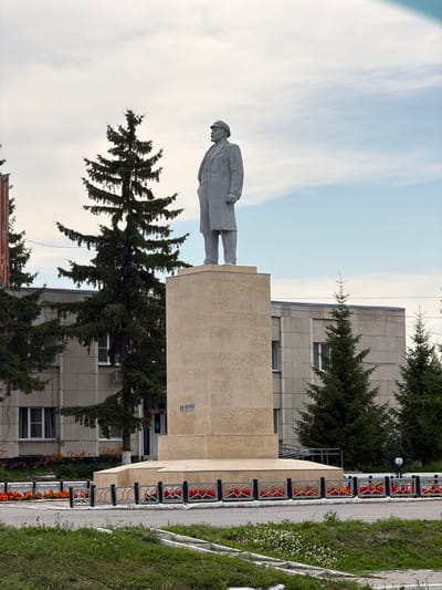 The image shows a statue of Vladimir Lenin in the center of Cherdakly, Russia. The statue is made of granite and depicts Lenin standing on a pedestal. The pedestal is inscribed with the words "В.И. ЛЕНИН" (V.I. Lenin). In the background, there is a modern building with several windows and a few evergreen trees. The statue is surrounded by a decorative fence and flower beds. The sky is overcast with some patches of blue.