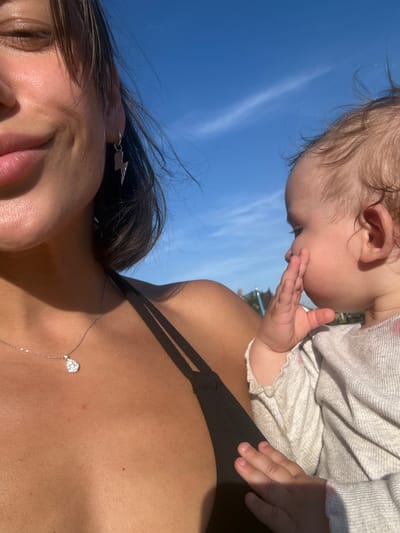 The image captures a close-up, low-angle shot of two individuals against a bright blue sky with wispy white clouds. The main subjects appear to be a woman and a baby. The woman is positioned in the foreground, with her face and upper chest visible. She has dark hair, some of which is blowing in the wind, and is wearing a black top. A delicate silver necklace with a teardrop-shaped pendant is visible on her chest. An arrow-shaped earring is also visible in her ear.
Behind her, and slightly above, is a baby. The baby's head and upper body are in view, with their head turned away from the camera, and their hand near their mouth. The baby is wearing a light-colored, long-sleeved garment. The overall impression is one of a sunny day, likely outdoors, given the clear blue sky and sunlight illuminating the subjects. The low angle suggests the photo was taken from the woman's perspective, perhaps by a third party or a selfie stick. No text is visible in the image. The setting is ambiguous beyond being outdoors under a clear sky.
