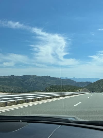 The image shows a view from inside a vehicle traveling on a highway. The highway is paved and has white lane markings. There are guardrails on the left side of the road, separating the highway from the surrounding terrain. In the distance, there are rolling hills covered in green vegetation and trees, with layers of mountains further back. The sky is blue with scattered white clouds. The location is specified as Pelev Brijeg, Montenegro.
