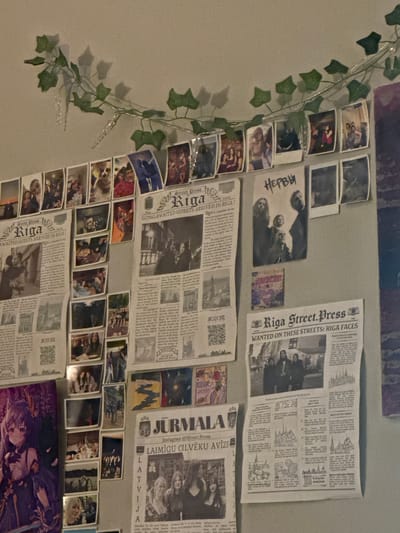 The image displays a wall decorated with various pieces of paper, predominantly newspapers and photographs, interspersed with a string of artificial ivy and lights. The newspapers appear to be from "Riga Street.Press" and "Jurmala," with headlines such as "LONG-AWAITED GUESTS ARRIVED IN RIGA!" and "LAIMIĞU CILVĒKU AVĪZE." The photographs are a mix of candid shots and posed portraits, featuring groups of people, individuals, and some scenic views. Some of the newspapers and photos are layered or overlapped. The overall impression is of a personal display or a collage, possibly in a bedroom or a common area. The location of Olaine, Latvia, is not visible or suggested by any elements within the image.