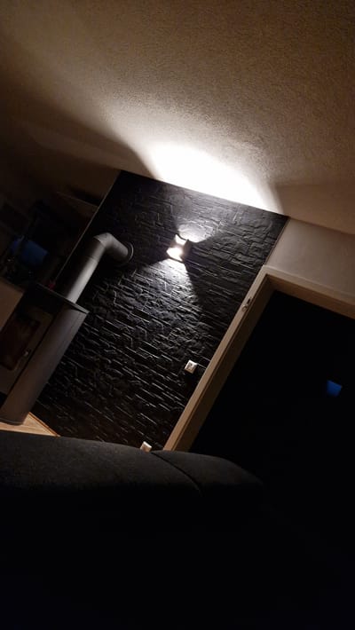 The image shows the interior of a dimly lit room, likely a living space, with a focus on a black stone-effect wall. A modern, dark gray pellet stove with a silver flue pipe is visible on the left, positioned against a light-colored floor. The wall itself is textured with horizontal stone-like tiles, and two wall-mounted lights cast a warm glow upwards and outwards, creating highlights and shadows on the textured surface. A white electrical outlet is visible on the wall below the lights. In the foreground, a dark upholstered piece of furniture, possibly a sofa, is present, though it is largely in shadow. To the right of the stone wall, a doorway or passage with a light-colored frame leads to a dark, indistinct area, possibly another room or the outdoors. The ceiling, also visible in the upper portion of the image, is textured and lit from above by an unseen light source, creating a soft, diffused illumination that contrasts with the direct lighting on the wall. The overall impression is one of a contemporary, cozy interior, possibly in a home in Ichenhausen, Germany, where the use of materials like stone and the atmospheric lighting contribute to a warm and inviting ambiance.