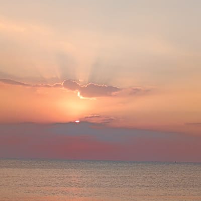 This image depicts a serene ocean sunset at Jangkar, Indonesia. The sun is partially obscured by soft, wispy clouds, casting a warm, golden-orange glow across the sky. Sun rays fan out from behind the clouds, creating a dramatic and ethereal effect. The lower portion of the sky transitions into softer pink and muted purple hues, suggesting the twilight hour.
The sea is calm, reflecting the colors of the sky in gentle ripples. A small, dark silhouette of a sailboat is visible on the horizon, adding a touch of scale and peaceful solitude to the vast expanse of water. The overall atmosphere is tranquil and contemplative, evoking a sense of peace and wonder as day transitions into night. The scene appears to be captured during a clear evening with fair weather. There is no visible text in the image.