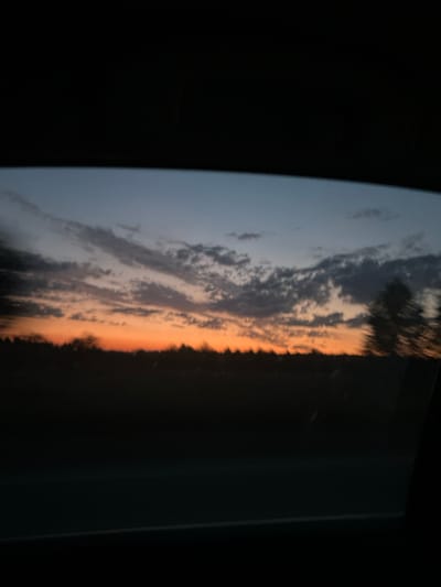 The image depicts a dusky sky viewed through a car window, with a dark silhouette of trees or a treeline in the distance. The sky exhibits a gradient of colors, transitioning from a deep orange near the horizon to lighter shades of blue and gray higher up. Wispy clouds are scattered across the sky. The foreground is dominated by the dark interior of the car, suggesting the photo was taken while in motion. There are no people or distinct objects visible, only the natural landscape and the car's interior framing the view. The setting appears to be rural or on the outskirts of a settlement, potentially in or near the village of Поварёнки, Russia, given the description.