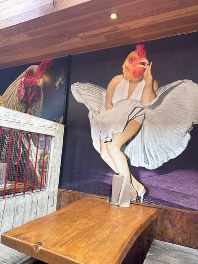 This image shows the interior of a space in Costa Mesa, United States of America, featuring eclectic decorations. On the wall are large posters: one of a woman in a Marilyn Monroe-esque pose, but with a chicken head, and another with a close-up of a rooster. A decorative repurposed wooden object, potentially an old door or window frame, with red-painted bars and metal mesh, is also on the wall. Part of a wooden countertop or table is visible, suggesting a commercial establishment like a restaurant or bar. There are no people visible in the scene.