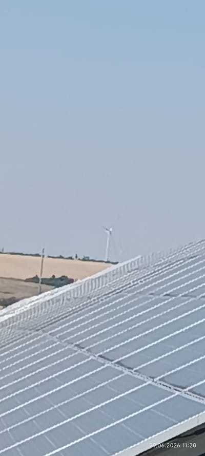 In Berezanka, Ukraine, on June 29, 2026, at 11:20 AM, a wide shot captures a bright, clear sky over a rural landscape. In the foreground, a large array of solar panels stretches across the frame, their metallic surfaces glinting. Beyond the panels, a distant view reveals a field of light brown earth, bordered by a darker green line of vegetation. A solitary wind turbine with three blades stands on the horizon, its structure thin against the sky. A few faint figures are visible in the distance, near the field, suggesting human presence in the area. The overall scene depicts a modern energy installation in a natural, open setting.