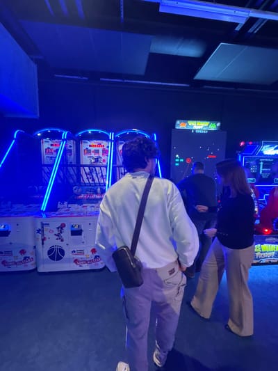 This image depicts an indoor arcade facility, likely located in Saint-André-lez-Lille, France, characterized by low blue ambient lighting. The scene features three individuals amidst various arcade game machines.
In the foreground, a person with dark, curly hair, viewed from behind, wears a white long-sleeved shirt, light-colored cargo pants, and white athletic shoes. A dark cross-body bag with a cylindrical pouch hangs from their left shoulder to their right hip. This individual stands facing towards the left-center of the frame, observing the arcade games.
To the right, a female with long, reddish-brown hair is partially visible, facing towards the left. She wears a dark long-sleeved top and wide-leg light-colored trousers with dark shoes. Her right arm is slightly raised.
In the background, a male figure is observed from behind, standing in front of an arcade machine. He wears a dark top and dark trousers and appears to be operating the game.
The environment is dominated by arcade machines. On the left, a series of three "Sonic All-Stars" basketball shooting games are prominently displayed, illuminated by bright blue neon lights framing the hoops. Score displays show "377", "1023", and "170". The machines feature "SEGA" and "SONIC SPORTS" branding. Directly in front of the male figure in the background, a "Space Invaders" arcade machine is visible, displaying the classic game screen with pixelated invaders, a player's ship, "1 CREDIT", and a score of "2000". To the far right, another blue neon-lit arcade machine is partially visible, displaying text that includes "HOW TO PLAY". The floor is a dark, uniform color, and the ceiling features exposed structural elements and acoustic panels, all under the pervasive blue light.