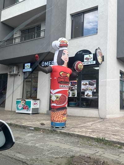The image captures a street view in the city of Aytos, Bulgaria, featuring a restaurant called "Ömeroglu Çiğköfte & Baklava". A prominent inflatable advertising figure of a woman in a chef's hat stands outside the establishment, holding what appears to be a plate of food. The restaurant's facade displays various promotional materials, including images of food items like çiğ köfte, baklava, and ice cream. An air conditioning unit is visible on the building wall to the left, and a refrigerator with "Medina Ice Cream" branding is placed on the sidewalk. The street appears to be paved with concrete slabs, and there is some debris on the ground, including a plastic bottle. A car's side mirror is partially visible in the foreground. The scene depicts a commercial area with typical urban elements.