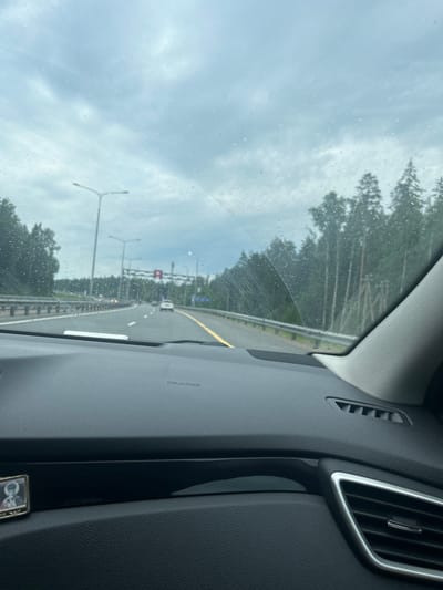 The image is taken from the interior of a vehicle, looking out through the windshield. The windshield has water droplets on it, indicating that it is raining or has recently rained. The view through the windshield shows a highway with multiple lanes. There are cars on the highway, some in the distance. Trees line the right side of the highway. There are streetlights and overhead signs visible. The dashboard of the car is in the foreground. On the left side of the dashboard, a small religious icon is attached. The sky is overcast and appears to be a pale blue-gray. The scene suggests a cloudy, possibly rainy day on a highway outside of a city.