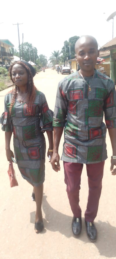 The image features a man and a woman walking side-by-side on an unpaved road in Ohafia, Nigeria, under bright daylight. Both individuals are dressed in coordinated outfits made from the same distinctive patterned fabric, which features a grid design of green, red, and grey squares with intricate motifs.
The woman, on the left, wears a knee-length dress with a V-neckline and ruffled, three-quarter sleeves. She carries a reddish-brown handbag in her left hand and wears black open-toed shoes. Her hair, which appears to be reddish-brown and long, is partially covered by a black head wrap. She also wears a silver necklace with a small pendant. Her gaze is directed slightly to her left.
The man, on the right, wears a matching tunic-style top over dark red or maroon trousers. He is bald and wears a gold chain with a small pendant, as well as a gold watch on his left wrist. His footwear consists of shiny black loafers with a gold emblem on the front. He looks directly towards the camera with a neutral expression.
The setting is an outdoor street scene, likely within a town or village. The road appears to be made of dirt or is unpaved. Buildings of various sizes, some multi-storied, line both sides of the road. Numerous trees, including palm trees, and utility poles are visible in the background. Further down the street, other people, cars, and motorcycles indicate a degree of activity. The bright sunlight casts noticeable shadows on the ground. There is no visible text in the image.