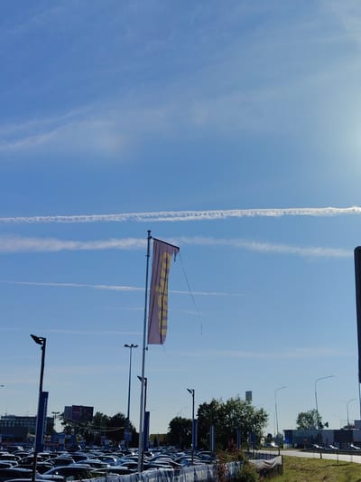 The image depicts an outdoor scene in Bielany Wrocławskie, Poland, likely a car dealership lot under a bright blue sky with contrails from airplanes. In the foreground, a large number of cars are parked in rows. A flag, which appears to be purple with yellow lettering, is prominently displayed on a flagpole in the center-right of the image, fluttering in the breeze. Several lampposts are also visible, as well as some trees and buildings in the background. The overall impression is of a commercial area on a clear day.