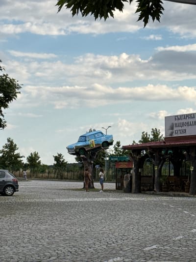 The image depicts an outdoor scene in Polsko Padarevo, Bulgaria, featuring a unique roadside attraction. A light blue car is perched precariously on a stylized tree structure, seemingly as a decorative element. The car is positioned at an angle, with its rear end higher than its front. It has several items visible inside, including what appear to be bottles.
Beneath the car and its tree support, there is a covered outdoor seating area with wooden pillars designed to resemble tree trunks. A sign with Bulgarian text, reading "БЪЛГАРСКА НАЦИОНАЛНА КУХНЯ" (Bulgarian National Cuisine), is visible above the structure.
In the foreground, a cobblestone paved area suggests a parking lot or walkway. A dark grey car is parked on the left side of the frame. Several individuals are present. A woman in a white shirt and blue shorts stands near the car on the tree structure. Another person, appearing to be a man with brown skin and wearing a brown garment, stands near the base of the tree support. Further in the background, a woman in a white t-shirt and denim shorts is visible, and another person is seen walking near a fence line.
The environment is characterized by greenery, with trees and shrubs lining the background. The sky is partly cloudy, with white cumulus clouds scattered across a pale blue expanse. The overall impression is a roadside establishment that uses an unusual display to attract attention.