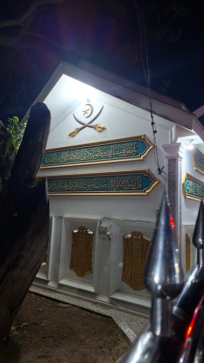 The image shows the facade of a building at night in Lhokdagang, Indonesia. The building is white with decorative elements in gold and teal. Above two horizontal bands of intricate Arabic calligraphy, there are two crossed swords with a crescent moon and star emblem. The calligraphy appears to be verses from the Quran. Below the calligraphy, there are decorative panels with a golden, lattice-like design set within teal borders. A large, rough-barked tree trunk is visible on the left side of the frame. In the foreground, out-of-focus, is a metal railing with pointed finials, some of which reflect a red light. The ground in front of the building is composed of dark soil with patches of sparse green grass and a decorative tiled pathway. Lighting on the building appears to be artificial, casting a bright glow. There are no people visible in the image.
