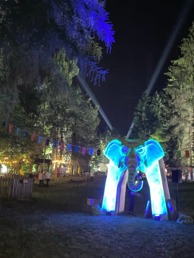 This nighttime scene, located in the Sremski Karlovci Municipality, Serbia, depicts an outdoor festival or event in a wooded environment. Central to the image is a large, illuminated archway shaped like a stylized elephant's head and trunk, glowing vibrantly in aqua blue and green. The word "playrix" is visibly inscribed in white on the elephant's forehead. One individual, dressed in dark clothing, stands underneath the archway, partially silhouetted. To the left, a string of colorful rectangular flags, displaying hues of blue, red, pink, purple, and green, extends across the upper mid-ground. Beneath these flags and behind a wooden picket fence, an area featuring numerous warm yellow string lights strung between trees is visible. Several indistinct figures, approximately three to five, are present in this background area, with two appearing to stand together. The surrounding tall trees are illuminated by various light sources; some branches in the upper left glow with a deep blue light, while others further back are bathed in warm, yellow tones. The dark night sky is bisected by two prominent white spotlights casting beams upwards from the mid-ground. A black speaker on a tripod stand is positioned to the right of the elephant archway. The immediate foreground consists of dark grass.