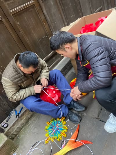 Two men are seated on the floor, likely working on decorations or lanterns. One man wears a black jacket and the other a beige quilted vest. Both are wearing blue jeans. They're surrounded by craft supplies including scissors, pliers, and colorful ornaments made of paper or fabric. One ornament appears to be red flower-shaped and another is yellow with red accents. A cardboard box sits nearby, seemingly containing more craft materials.