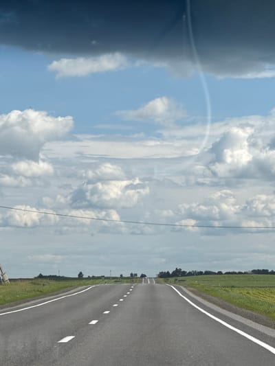 This is an outdoor, eye-level, daylight, wide-angle shot of a paved road in the city of Акцябрскі сельскі Савет, Belarus. The road has white dashed lines on the right side, indicating the edge of the lane, and solid white lines on the left. The road stretches into the distance, curving slightly to the right. On either side of the road are fields of green crops. The sky is filled with large, puffy white clouds and some darker gray clouds. In the far distance, there are trees and what appear to be cell towers. There is a thin, curved streak of light or reflection visible in the upper right portion of the sky. No people are visible in the image.