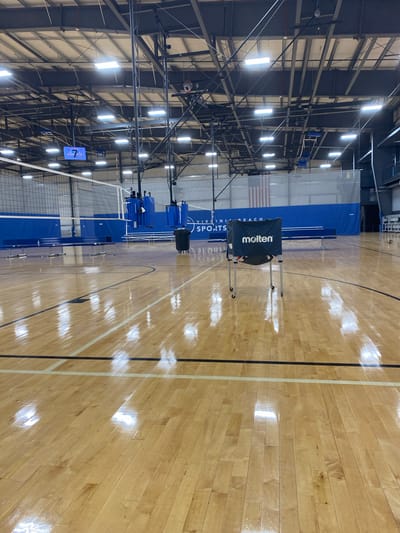 The image shows an indoor volleyball court. The court is made of polished wood flooring with black and white lines marking the boundaries. A volleyball net is set up in the background. There are blue padded protectors on the net poles, and a blue banner with "VIRGINIA BEACH SPORTS" written on it is visible behind the net. A black trash can on wheels is located to the right of the net. In the foreground, a portable ball cart with the "Molten" logo is positioned in the center of the court. The ceiling of the facility features exposed metal beams and industrial lighting. The overall environment appears to be a sports complex or gymnasium.