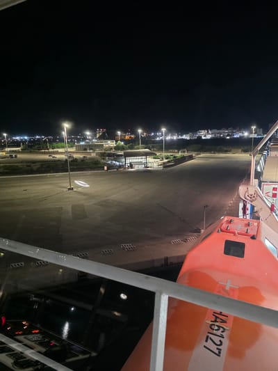 A night-time aerial view shows an empty industrial port area in Brindisi, Italy, with a large orange lifeboat visible in the foreground on the right. The lifeboat, marked with "JA6127" and a white cross, is positioned on a ship, partially obscuring the view of the port. The port area features paved surfaces, parked vehicles, streetlights illuminating rows of lights in the distance, and buildings with lit windows. The water is visible in the lower left portion of the frame.