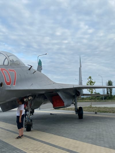 In Arablyar, Russia, a young person wearing a white t-shirt and dark shorts stands next to the landing gear of a grey military jet with the number "07" in red on its nose. The jet is parked on a paved surface with a patterned design of grey and yellow bricks. The sky is overcast with light grey clouds. In the background, there is a road, a grassy area with trees, and a few tall poles with lights. The scene suggests an outdoor display or museum exhibit.
