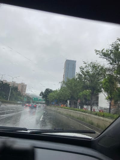 The image depicts a view from inside a vehicle traveling on a multi-lane road during a heavy rainstorm in Luohu District, China. The windshield is covered with raindrops, obscuring some details of the external environment. The road surface is wet and reflects the overcast sky and the headlights of other vehicles. Several cars are visible on the road, some with their brake lights illuminated, indicating slow or stopped traffic. On the left side of the road, there are apartment buildings and green trees. On the right side, there is a concrete barrier with more trees and what appears to be construction in progress, indicated by scaffolding and some incomplete structures. A tall, light blue building with multiple windows stands prominently in the background, partially obscured by trees. The overall impression is of a typical urban scene during inclement weather.