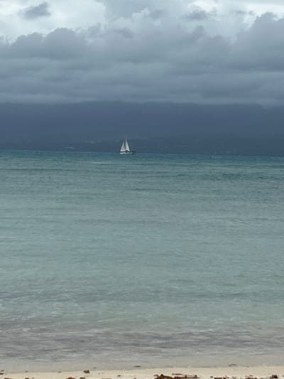 A horizontal landscape image shows a beach scene in Le Gosier, France. On the left side of the image, storm clouds dominate the sky above the ocean. The ocean itself is a gradient of deep gray-blue on the left to a lighter turquoise towards the beach on the right. A small white sailboat is located near the middle of the image. A sandy beach is visible along the right edge of the image.