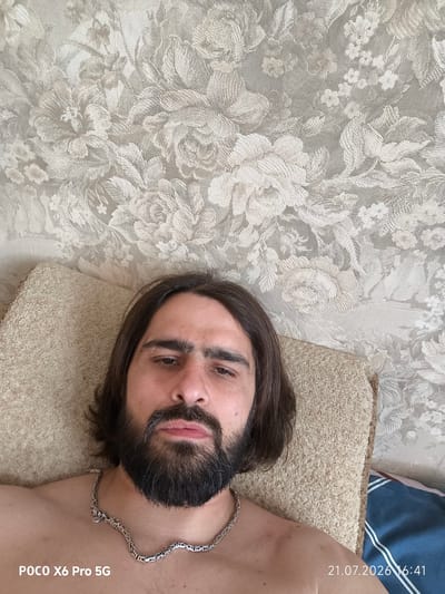 A man with long brown hair and a beard is lying on a pillow against a floral wallpaper background. He is wearing a silver chain necklace. The timestamp "21.07.2026 16:41" and "POCO X6 Pro 5G" are visible in the lower right and left corners of the image, respectively. The image appears to have been taken indoors.