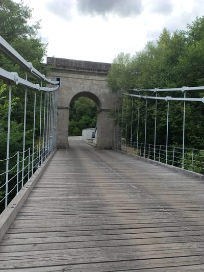 The image shows a perspective view of a wooden suspension bridge in Stádlec, Czechia. The bridge has a weathered wooden deck and white metal railings supported by vertical posts. Beyond the railings, dense green trees line both sides of the bridge. In the distance, an arched stone structure forms an entrance or gateway. The sky is overcast with heavy clouds. The scene is static, with no people or actions visible.