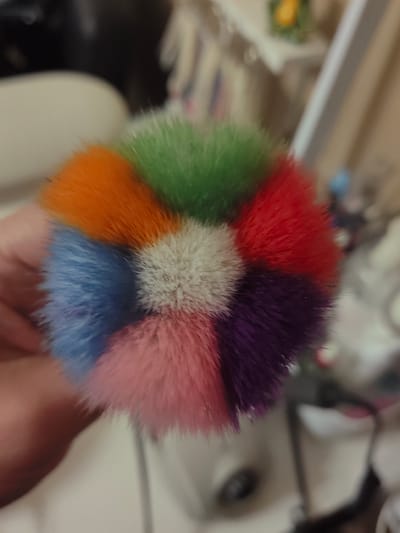 The image displays a close-up of a makeup brush with a multicolored head, featuring bristles in shades of orange, green, red, purple, pink, blue, and white. The brush head is held by a person's hand, part of which is visible in the lower left of the frame. The background is out of focus, showing what appears to be an indoor setting with some shelves and objects. There is no indication of the city of Ostafievo, Russia, in the image or its context.
