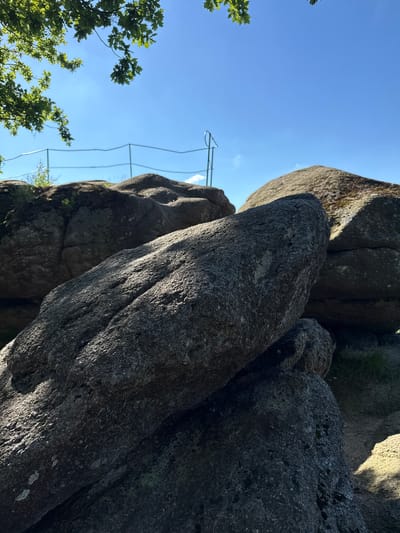 The image depicts a sunny outdoor scene featuring large granite boulders. In the foreground, a prominent boulder is illuminated by sunlight, highlighting its rough texture. Behind it, several other boulders are visible, with some partially in shadow. A thin metal railing with posts can be seen in the background, suggesting a viewing area or a pathway along the rocks. Green leaves from an overhead tree branch are visible in the upper left corner, with a clear blue sky filling the upper portion of the frame. The overall environment suggests a natural park or a scenic viewpoint. This is likely the "Pajęcza Baszta" (Spider Tower) rock formation in Szklarska Poręba, Poland, a popular tourist attraction known for its unique geological formations and views.