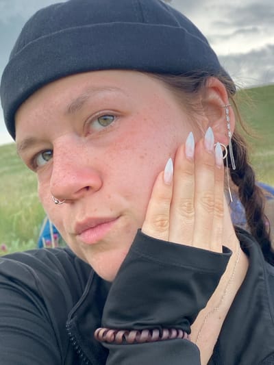A close-up portrait of a young woman wearing a black beanie and black jacket. She has light skin with freckles, green eyes, and her left hand is resting on her cheek. She is wearing a silver hoop nose ring and a silver earring with a chain. Her fingernails are long and painted white. A brown spiral hair tie is visible on her left wrist. The background appears to be an outdoor grassy area with a cloudy sky. The scene is likely in a rural setting, possibly near the village of Ziyanchurino, Russia, given the natural landscape.