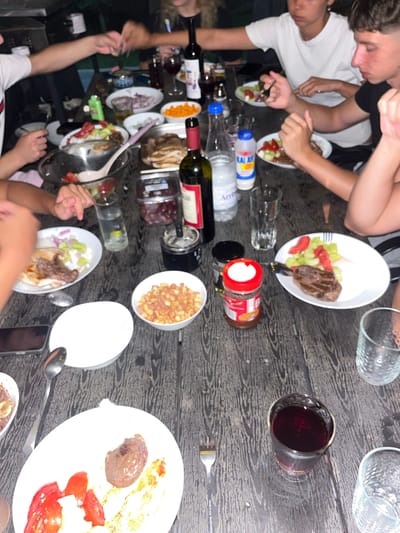 In Chaidefto, Greece, a group of people are gathered around a dark wooden table, sharing a meal outdoors. The scene is captured from a high angle, looking down at the table laden with food and drinks. Plates of steak with salad, bowls of what appears to be corn or beans, and bottles of wine are spread across the surface. Some individuals are visible, with their arms and hands reaching for food or holding utensils. The overall atmosphere seems casual and social, suggesting a relaxed gathering. The lighting suggests it might be evening or a dimly lit area. There are no strong indicators of the weather. Some text is visible on the bottles, including "KALAS" and "Arren" on what appear to be water bottles. A wine glass filled with red wine is prominently placed in the center of the frame.