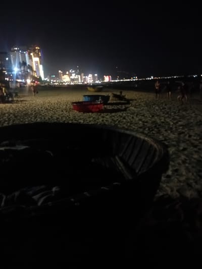 In Đà Nẵng, Vietnam, at night, the scene is a sandy beach with a city skyline in the background. The foreground is dominated by a dark, round boat, possibly a coracle, that is mostly out of focus. Beyond the boat, the sand is textured and populated by several other objects and people.
Two colorful bowls, one red and one blue, sit on the sand to the left of the center. Further back, more people are visible, some appearing to be walking or standing. One individual is partially obscured by shadow on the left. In the mid-ground, a person is walking towards the right.
The city skyline is illuminated by numerous lights, featuring tall buildings with glowing windows. The sky is dark, indicating it is nighttime. The overall impression is a beach scene at night with urban activity visible in the distance.