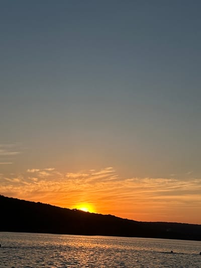 This image depicts a sunset scene at Ustia, Moldova. The sun is low on the horizon, casting a warm orange glow across the sky and illuminating wispy clouds with streaks of yellow. The silhouette of a dark hillside is visible against the vibrant sky. To the right of the hillside, a body of water, likely the Nistru River, reflects the sunset's colors, creating shimmering patterns on its surface. The water appears slightly choppy, with a few dark, circular objects floating on it, possibly buoys or markers. The overall mood is serene and peaceful, characteristic of a sunset over a natural landscape. The time of day is clearly evening, as indicated by the setting sun. No people or text are visible in the image.