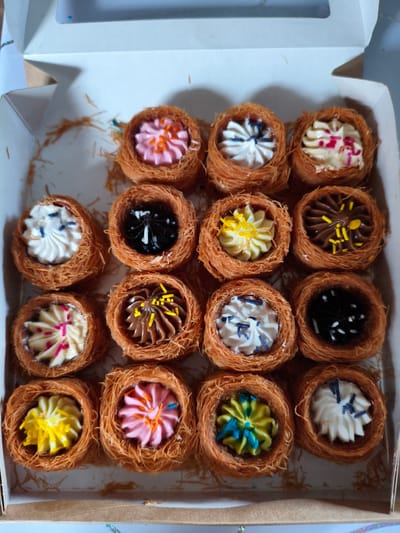 A box of 16 individual pastries sits inside a white cardboard box. Each pastry features a nest-like base of shredded filo dough, filled with a rosette of brightly colored cream. Cream colors include white, pink, yellow, chocolate, black, and a combination of yellow and green. Some creams are topped with colorful sprinkles. The location is presumed to be Мунайка, Russia, but there is no definitive evidence in the image to support this.