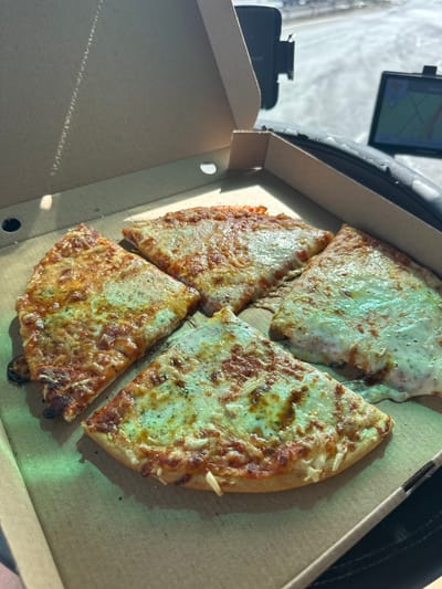 An overhead shot shows four slices of cheese pizza in a cardboard pizza box, bathed in sunlight. The pizza box is open and rests on a dark surface, possibly a car dashboard. To the right, a GPS device is visible, displaying a map with a route. The background is blurred, but appears to be an outdoor setting, possibly a street or road. The pizza slices are triangular and topped with melted cheese and herbs.
