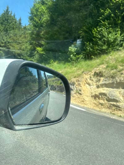 The image shows a view from inside a moving vehicle, looking out the side window and into the side-view mirror. The side-view mirror reflects the road behind the vehicle, lined with trees and a rocky embankment. The surrounding environment appears to be a rural, mountainous area with dense green trees and a clear blue sky. The road is paved and has a white line marking the edge. The vehicle itself is silver. The location is identified as Lavans-lès-Saint-Claude, France, suggesting it is a scenic route within the Jura Mountains. No people are visible in the image.