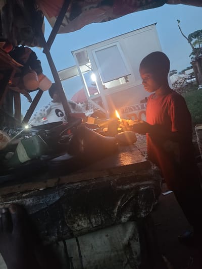 The image captures a dimly lit scene, possibly at dusk or night, with a young person in the foreground holding a lit candle. The person, wearing a red shirt, is focused on the flame. The background shows a white structure with some lighting, and elements that suggest an outdoor or makeshift setting. There are also hints of other people and objects, including what appears to be part of a person's face with a white and black patterned covering, and various items that look like containers or supplies.
The overall atmosphere is somewhat somber, possibly due to the low light and the individual's concentrated expression. There is visible text on what appears to be a white sign or advertisement in the background, reading "BENNY CO". The scene seems to be in Karu, Nigeria, given the provided location context. The image is oriented vertically, adding to its unique perspective. The presence of the candle suggests a need for light in the absence of sufficient illumination, possibly indicating a power outage or a remote location.