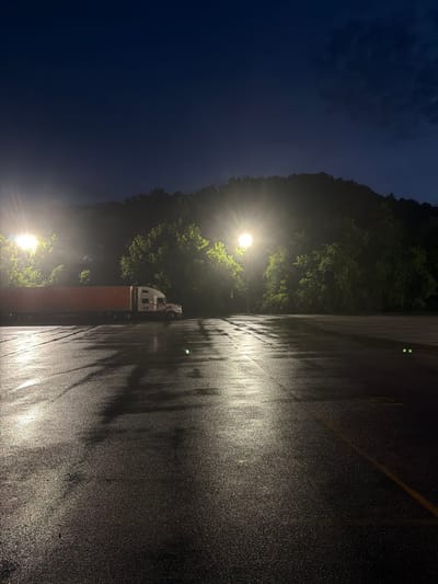 A semi-trailer truck is parked in a wet, empty parking lot at night. Two tall light poles illuminate the scene, casting long reflections on the dark, wet asphalt. Trees and a dimly lit hillside form the background. The sky is a deep blue, transitioning to black at the top of the frame. The truck is white and orange, with the trailer appearing to be a deep red or orange color. No people are visible in the image. The overall impression is one of stillness and isolation. The location is an outdoor parking lot in Wildwood, United States.