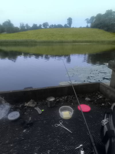 The image depicts a fishing scene at a body of water, likely a canal or river, with a grassy embankment on the far side. In the foreground, on what appears to be a wooden platform or bank, are various fishing accessories. These include a fishing rod, a small tin can, a clear plastic container with some yellow substance inside, a red lid, and a pair of scissors. The water surface is calm, with lily pads visible in the mid-ground. The overall atmosphere is overcast or hazy, suggesting it might be early morning or a cloudy day. There are no people visible in the frame. The location is identified as Roslea / Rosslea, United Kingdom.